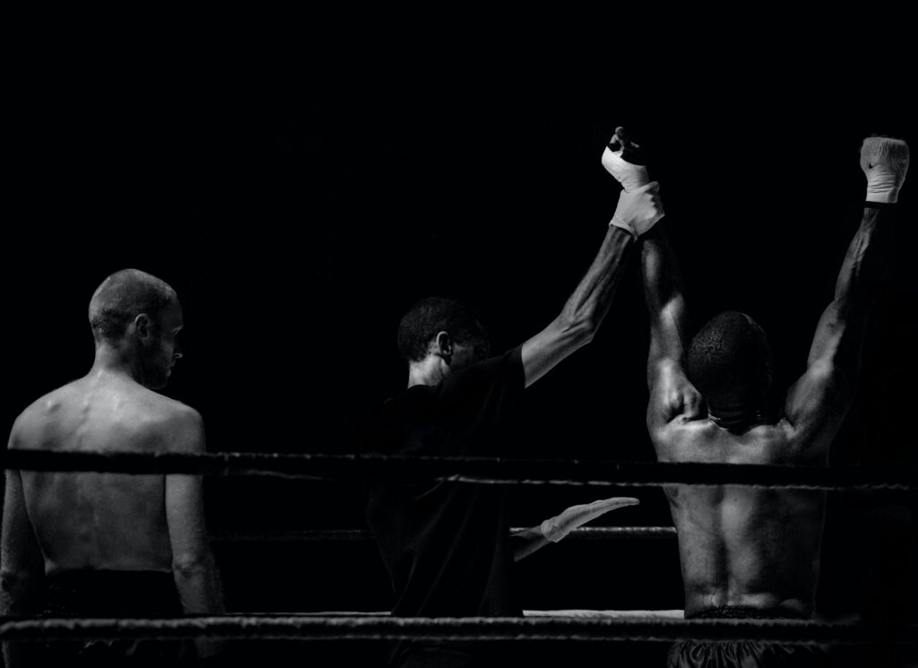 Photo taken from behind at the end of a bout where the ref lifts one of the boxers hands while the other boxer looks on.